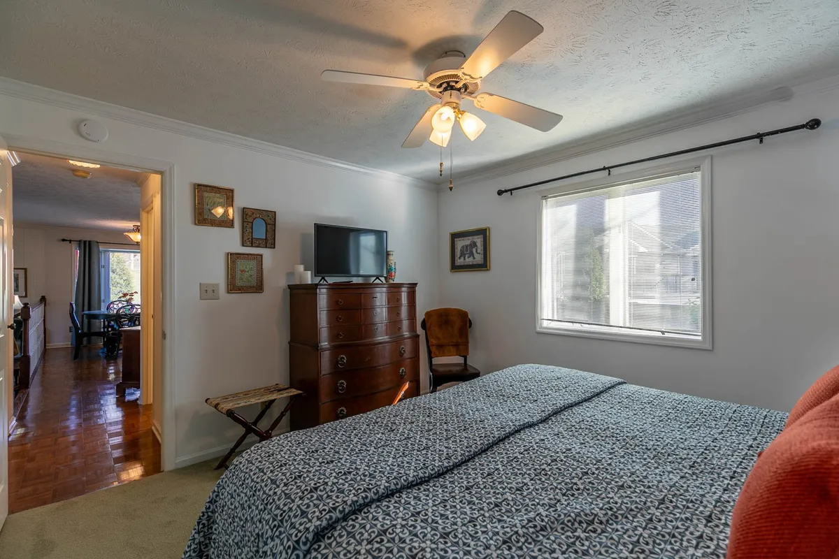 A bright bedroom with a patterned bedspread, a dresser topped by a television, and a comfortable reading chair near a window that lets in abundant natural light. The setup is perfect for morning awakenings or winding down with a show.