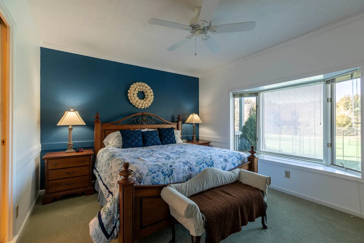 A serene bedroom featuring a cozy bed with floral bedding, a blue accent wall, and a bay window that fills the room with natural light. The view through the window emphasizes privacy and a connection to beautiful natural surroundings, ideal for rest and contemplation.
