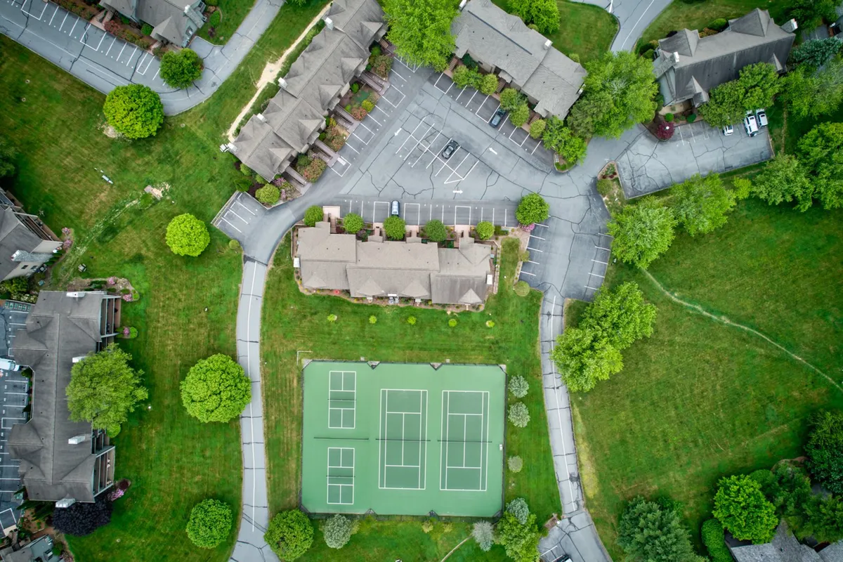 A panoramic view encompassing the tennis courts, surrounding greenery, parking area, and glimpses of residential buildings. The scene conveys a relaxed, social atmosphere with spaces for both active recreation and quiet strolls.