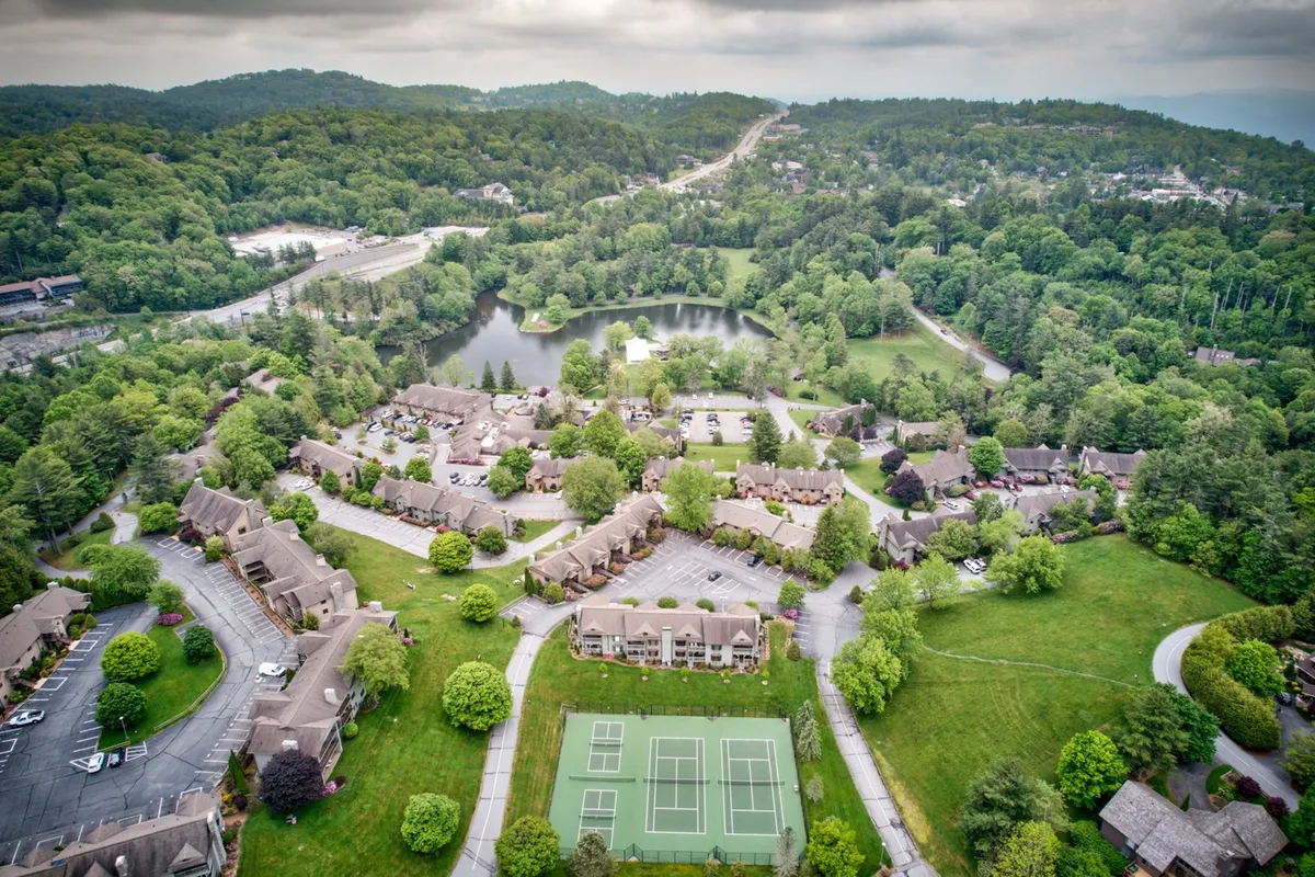An exterior view featuring lush greenery, a serene pond, tennis courts, and inviting townhouse-style accommodations. The setting exudes peacefulness, with open areas for walking, relaxing by the water, or enjoying an active afternoon.