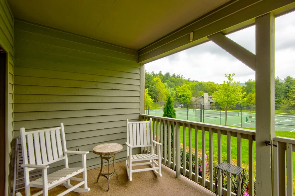 An inviting exterior space featuring rocking chairs positioned for watching tennis matches, with a small table and tennis courts in the background. The verdant landscape adds to the relaxed, spectator-friendly setting.