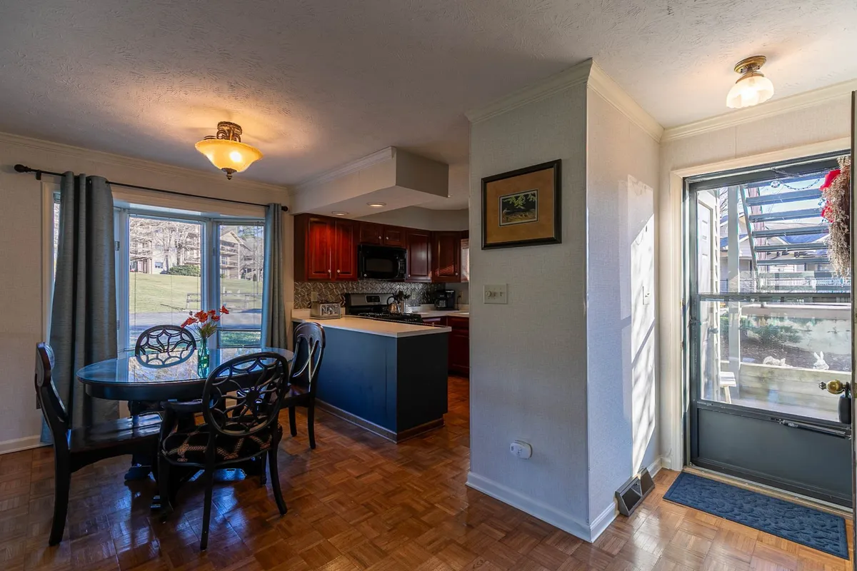 This image displays a warm and welcoming dining area with a sturdy wooden dining table, large windows letting in natural light, and a visible kitchen island in the background. The decor is tasteful and inviting, creating a cozy space perfect for meals or entertaining.