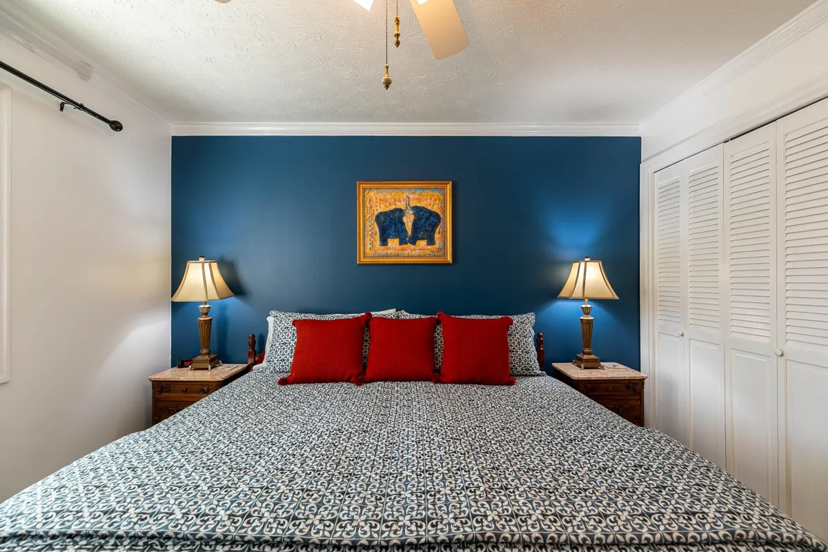 A cozy bedroom featuring a bed with patterned bedding, vibrant red accent pillows, and framed elephant artwork above. Soft table lamps flank the bed, casting a warm glow, and the intimate space invites relaxation and restful sleep.