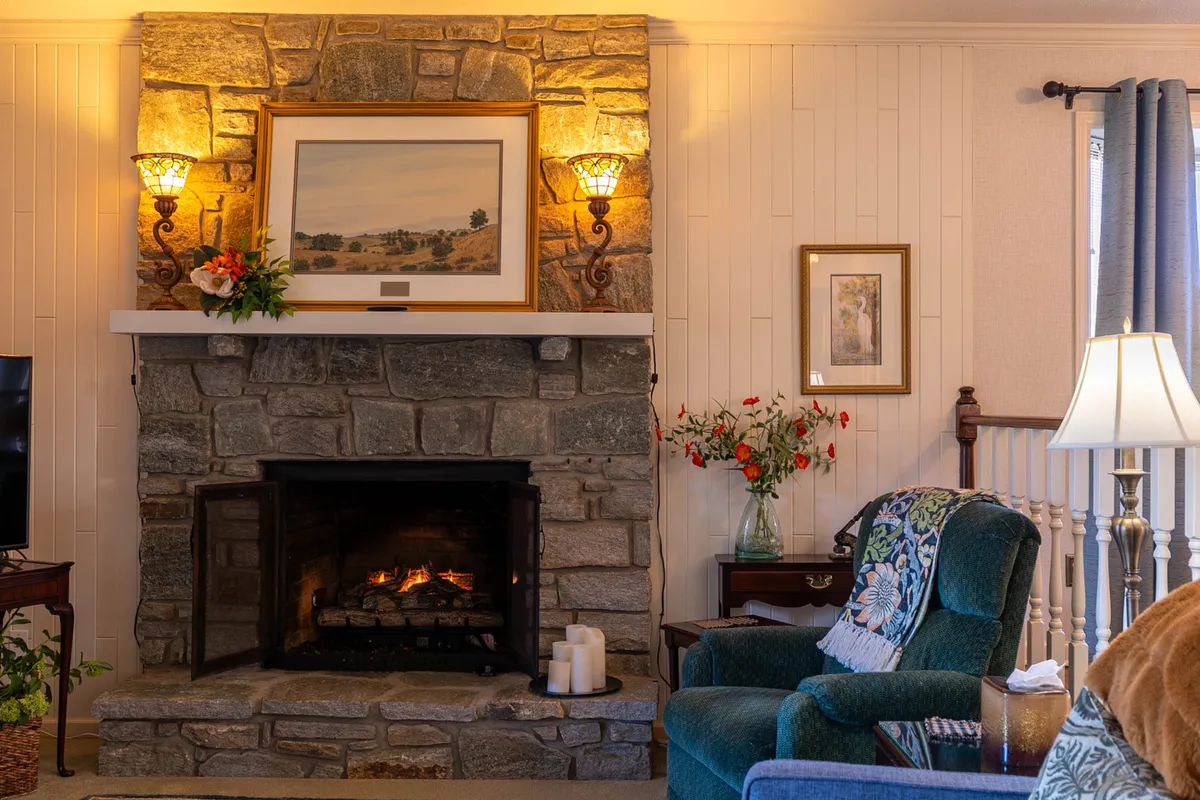 A cozy nook highlighted by a stone fireplace, green armchair, table lamp, and tasteful wall art. The intimate setting is ideal for curling up with a book or enjoying a quiet moment by the fire.